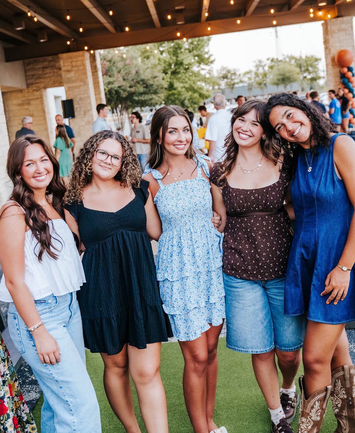 Five Austin Christian University students standing arm in arm, smiling, under string lights at an outdoor evening event on campus.